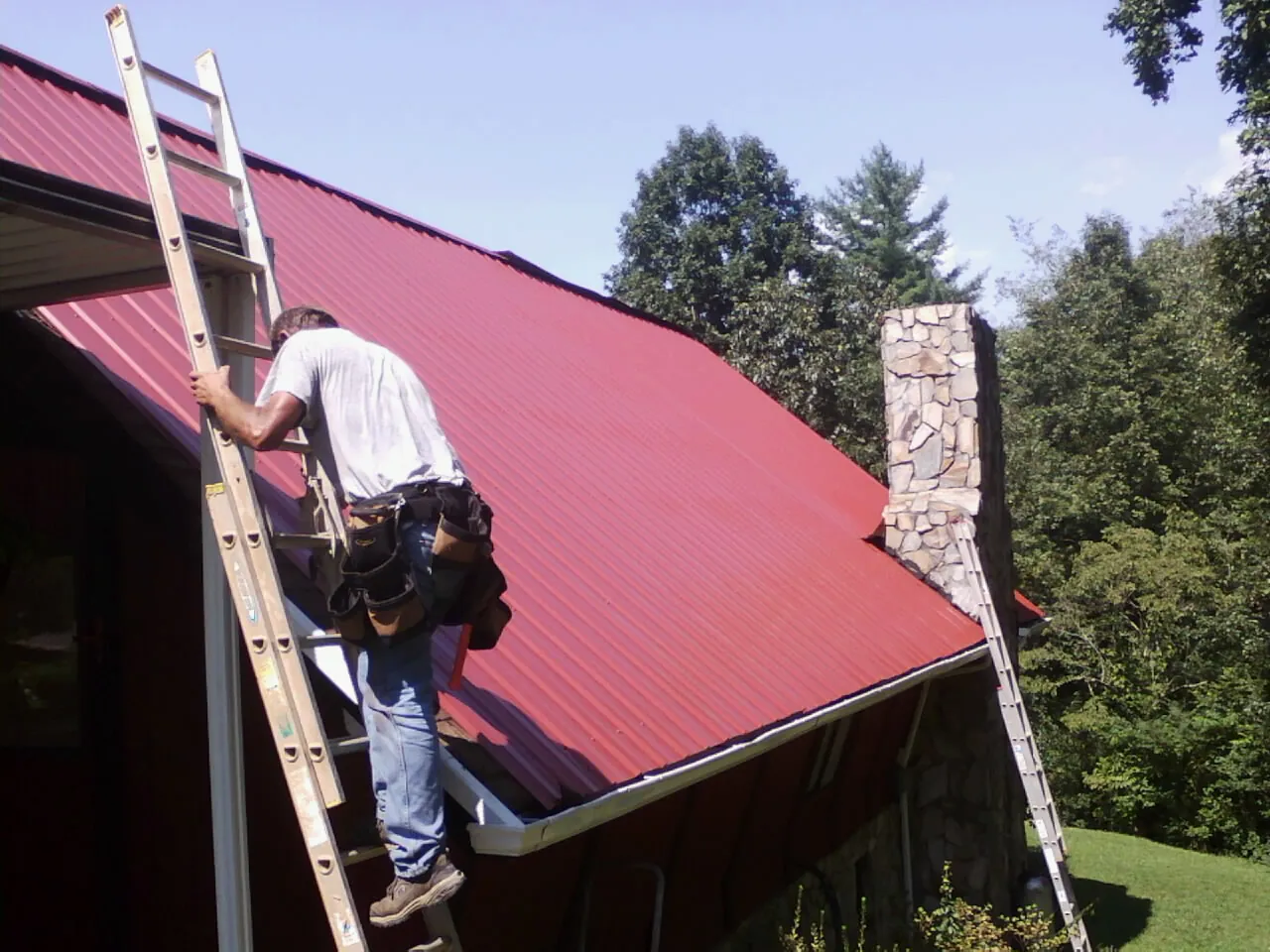 Roofer on a ladder beside a red metal roof and stone chimney — Soffit & Fascia Repair by Roofing Brothers in Bismarck